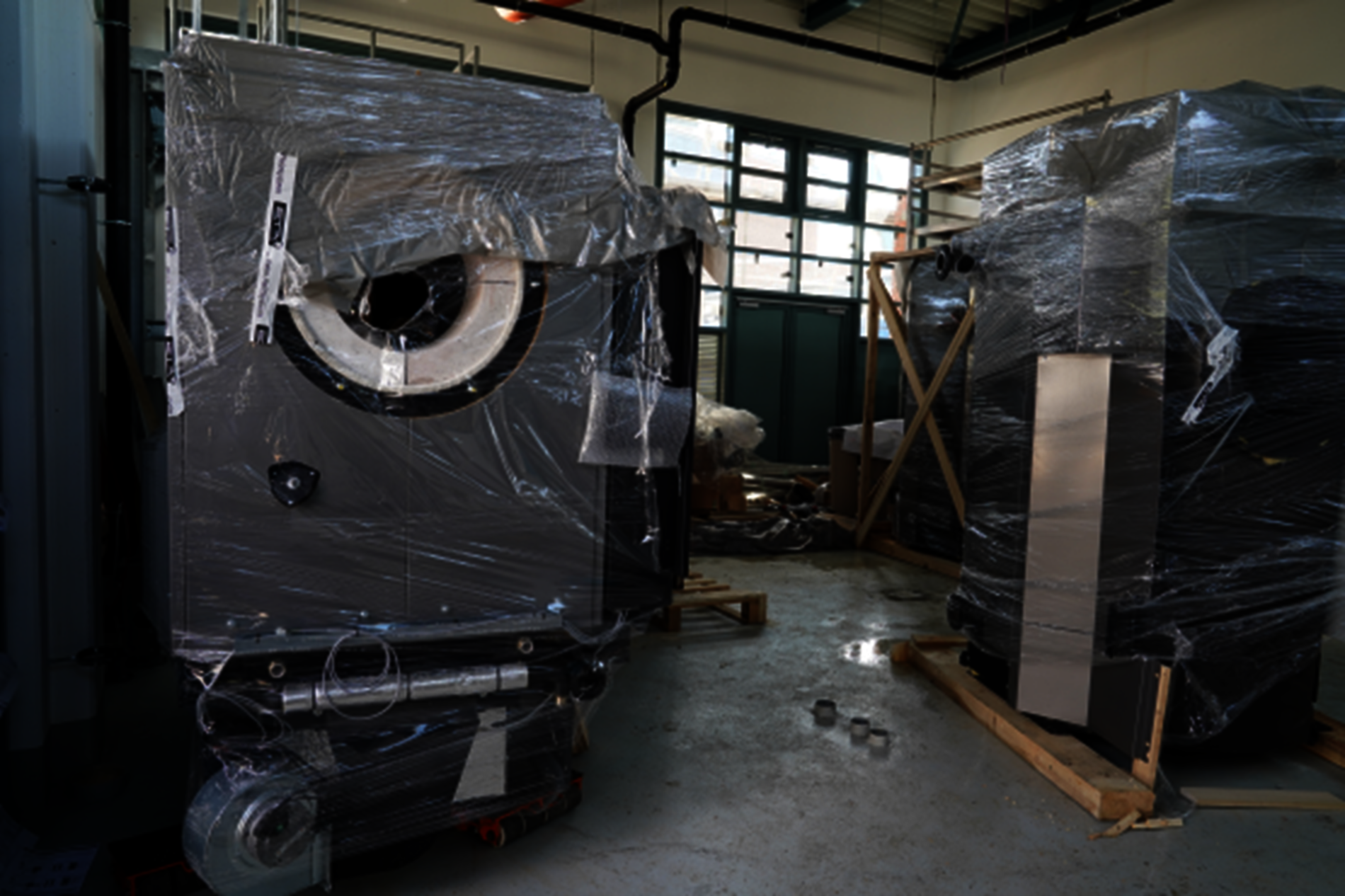 Zwei große graue Heizkessel stehen in Plastikfolie verpackt in einer Halle.