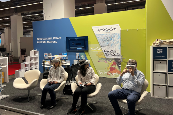 Drei Personen mit weißen VR-Brillen sitzen in Stühlen am Messestand der BGE auf der Buchmesse in Frankfurt am Main.