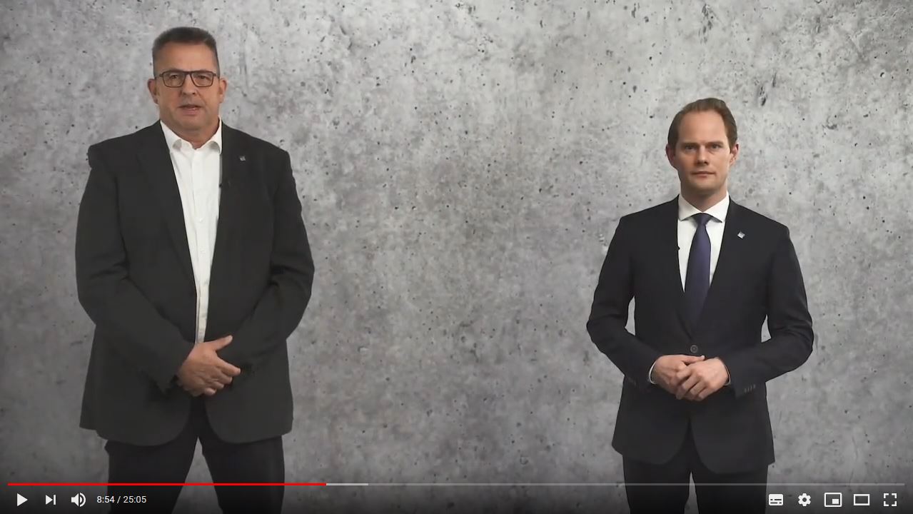 Zwei Männer in Anzügen stehen vor einer grauen Wand