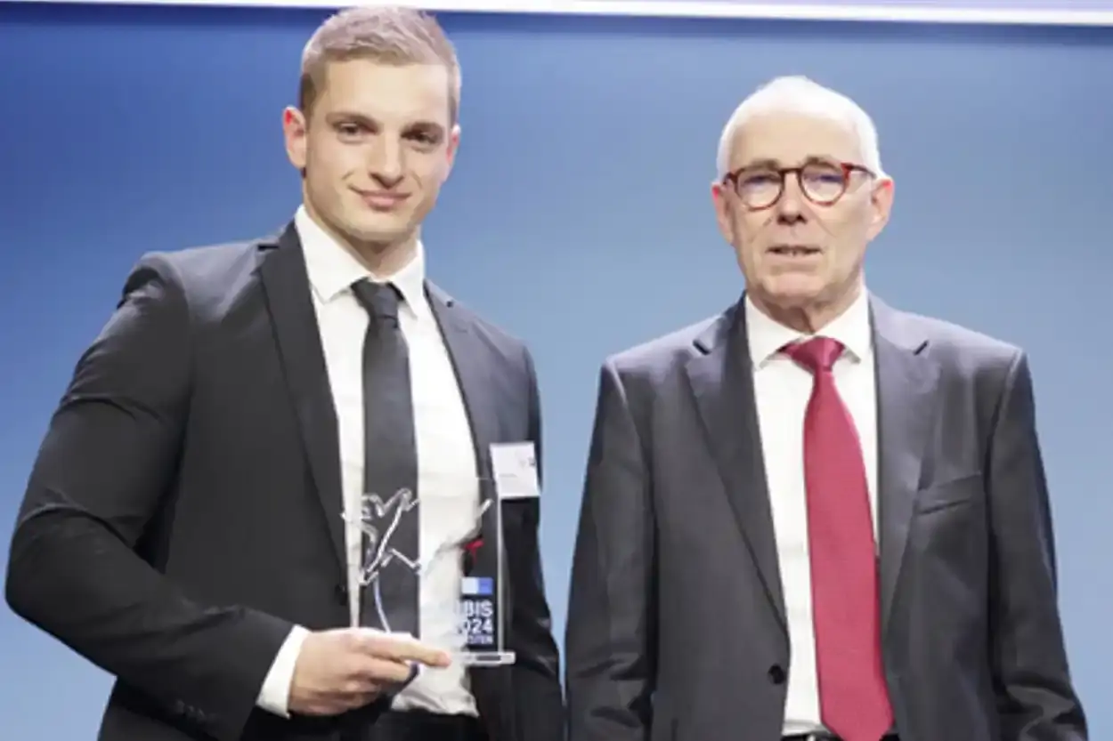 Das Foto zeigt zwei Männer in dunklen Anzügen und weißen Hemden. Der linke Mann hält eine halbtransparente Trophäe in seiner rechten Hand und lächelt in die Kamera.