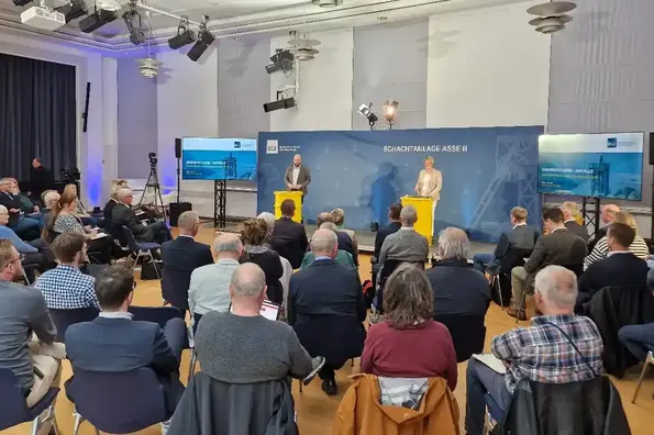 Zu sehen ist ein großer Raum. Im Hintergrund befindet sich eine Bühne. Auf der Bühne stehen ein Mann und eine Frau an gelben Fässern, die als Tisch benutzt werden. Im Vordergrund sitzen Menschen auf Stühlen und hören den Vortragenden zu.