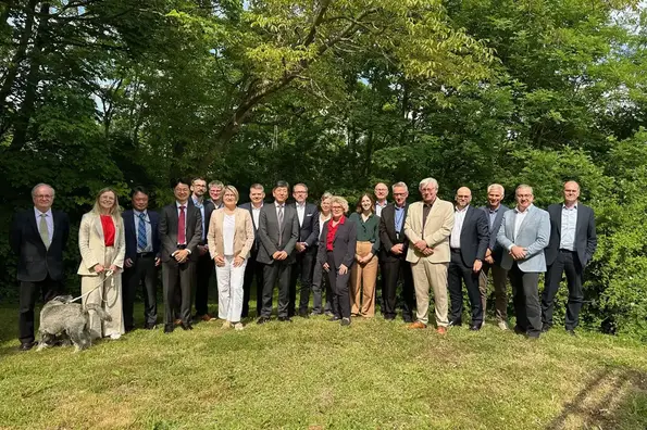 Gruppenfoto von 20 Personen, die bei frühlingshaftem Wetter auf einer Wiese vor Bäumen mit grünen Blättern stehen. Eine Frau hält einen Hund an einer Leine.