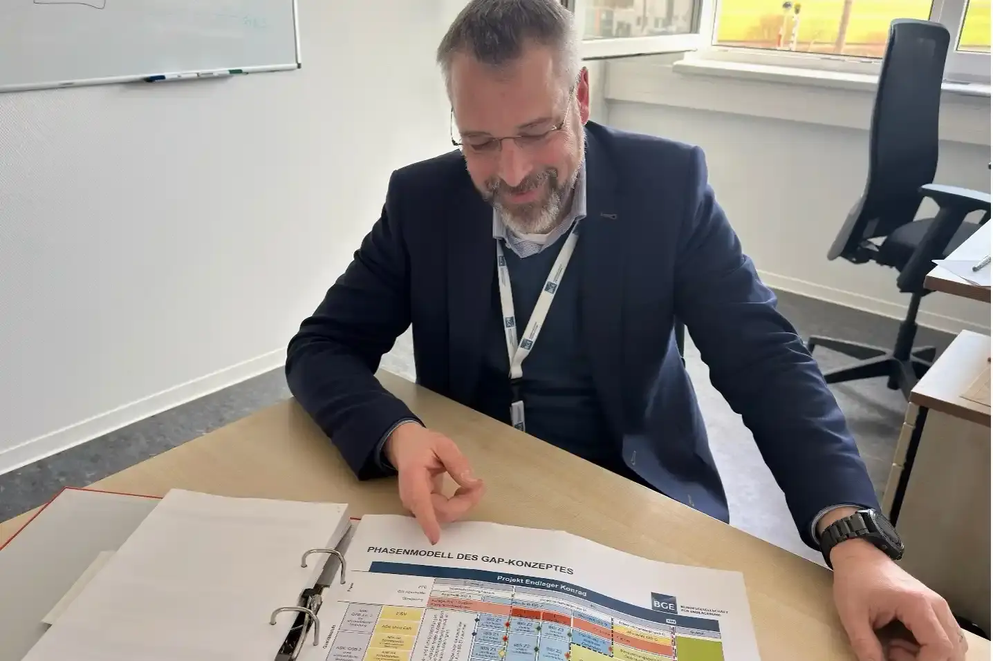 Ein geöffneter Ordner mit Planungsunterlagen liegt auf einem Schreibtisch. Ein Mitarbeiter mit Vollbart und Brille sitzt auf einem Stuhl und zeigt auf den geöffneten Ordner.