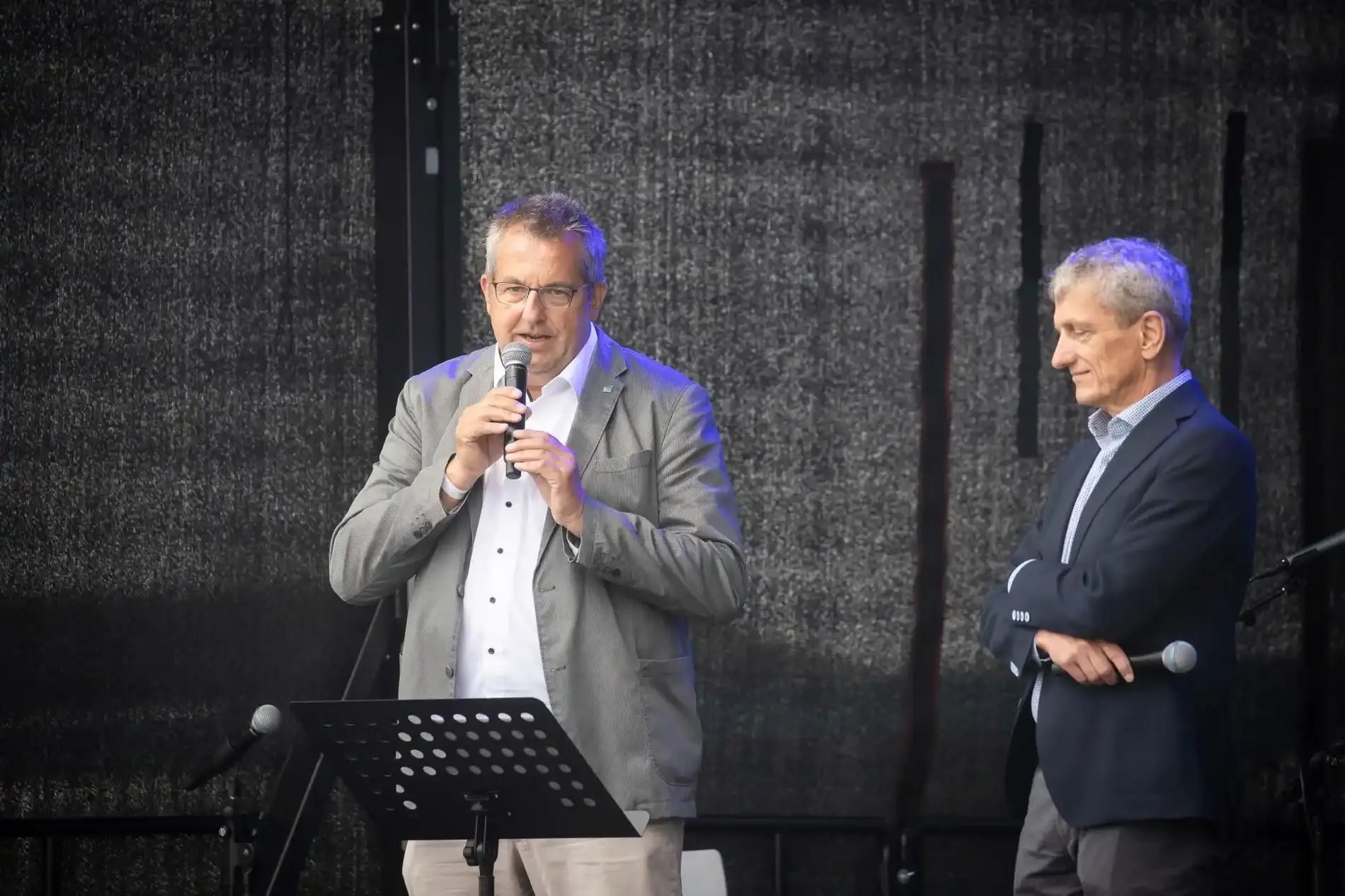 Zwei Männer in Anzug stehen auf einer Bühne. Einer spricht in ein Mikrofon.