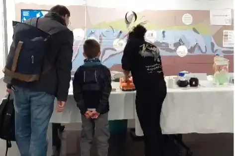 Zwei Erwachsene und ein Kind beugen sich über einen Tisch, auf dem verschiedene Gegenstände liegen. Im Hintergrund sind verschiedene geologische Grafiken erkennbar.