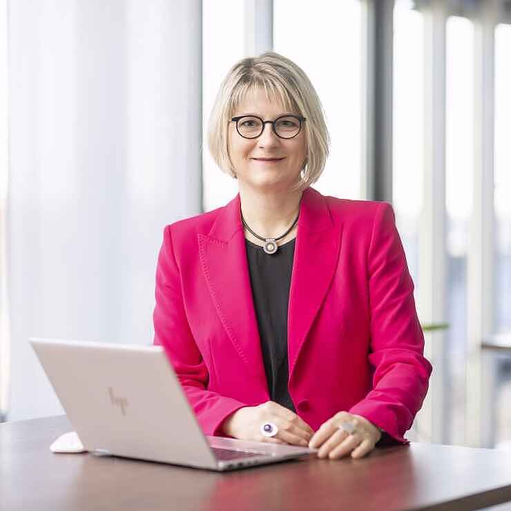 Eine Frau mit kinnlangen blonden Haaren und Brille steht an einem Stehtisch. Sie trägt einen pinken Blazer und darunter ein schwarzes Oberteil. Vor ihr auf dem Tisch steht ein aufgeklapptes Notebook. 