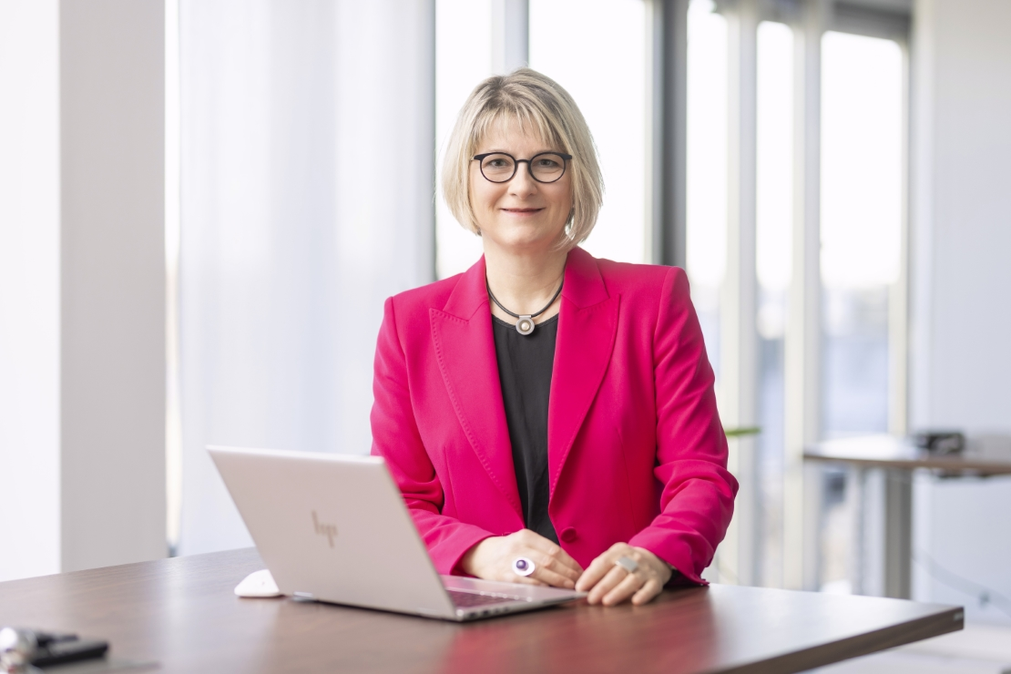 Eine Frau mit kinnlangen blonden Haaren und Brille steht an einem Stehtisch. Sie trägt einen pinken Blazer und darunter ein schwarzes Oberteil. Vor ihr auf dem Tisch steht ein aufgeklapptes Notebook. 