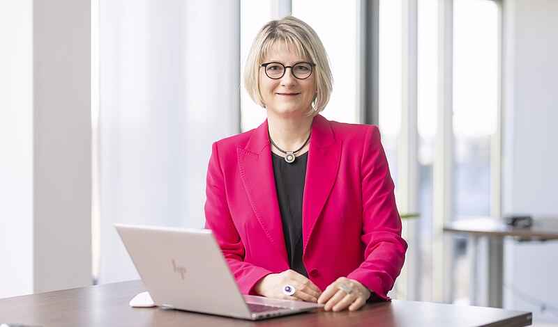 Eine Frau mit kinnlangen blonden Haaren und Brille steht an einem Stehtisch. Sie trägt einen pinken Blazer und darunter ein schwarzes Oberteil. Vor ihr auf dem Tisch steht ein aufgeklapptes Notebook. 