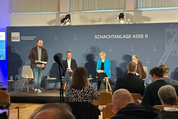 Eine stehende Person und drei sitzende Personen vor einem gut gefüllten Saal.