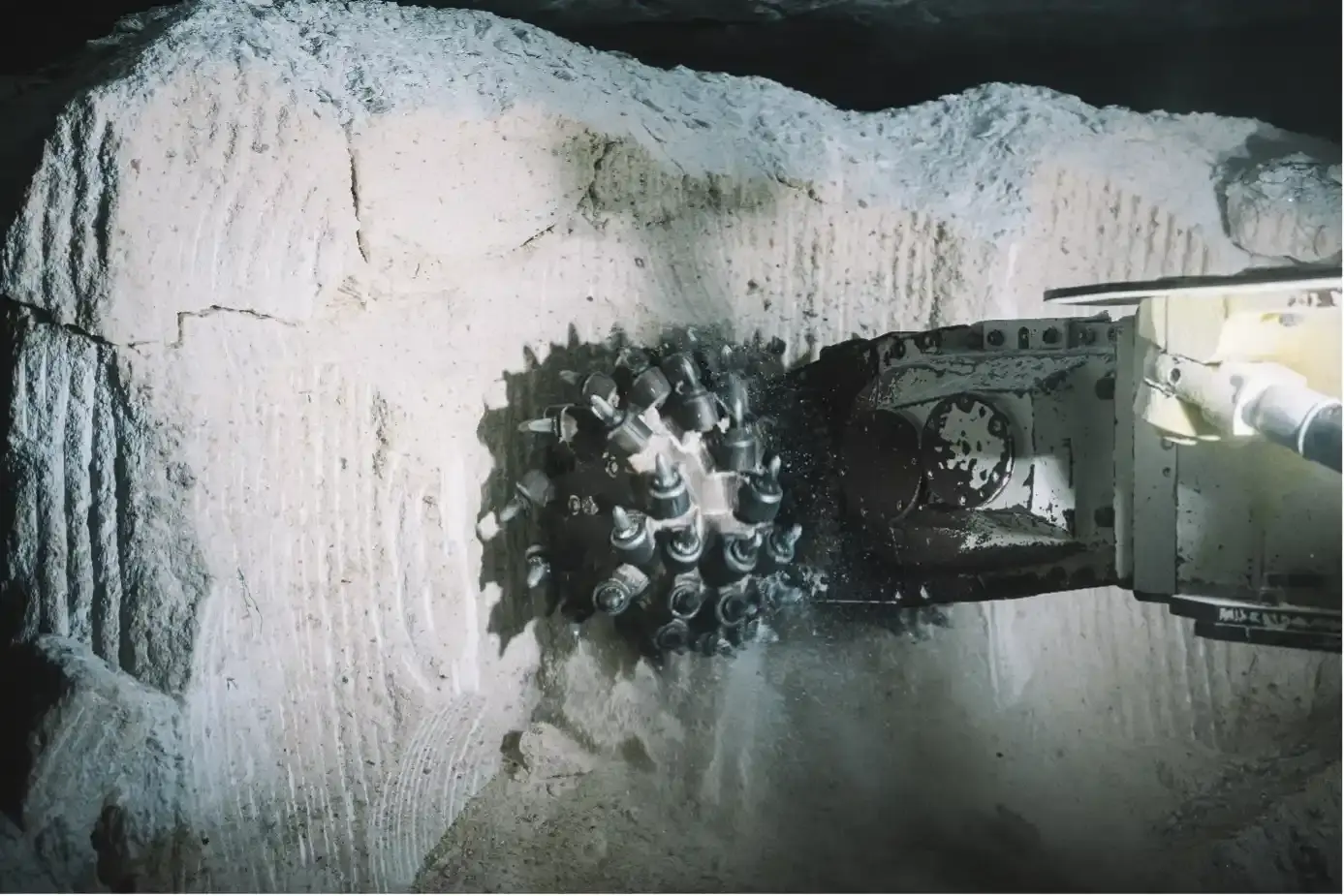 Der drehende Schneidkopf einer Teilschnittmaschine fräst sich in Salzgestein und hinterlässt dort linienförmige Strukturen. Das abgefräste Material fällt in einer kleinen Staubwolke auf den Boden des Bergwerks.