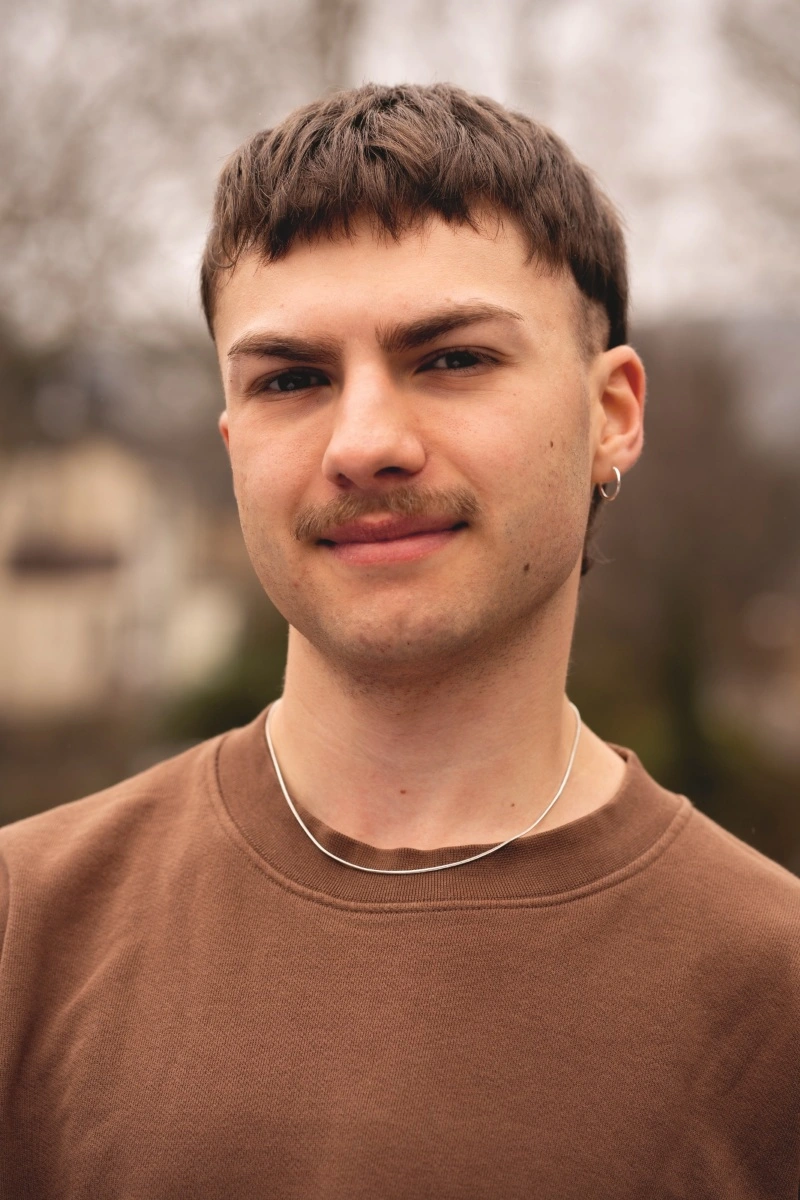 Portrait eines jungen Mannes mit braunen kurzen Haaren und Oberlippenbart. Er trägt eine silberne Kette sowie einen Ohrring und einen braunen Pullover.