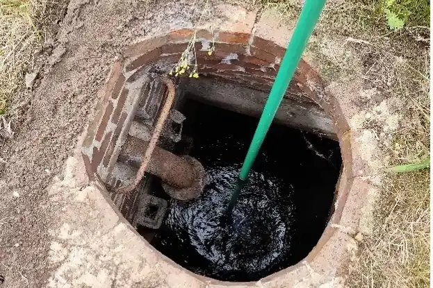 Blick von oben in eine mit Wasser gefüllte Zisterne. Eine Hand hält einen grünen Stab ins Wasser. 