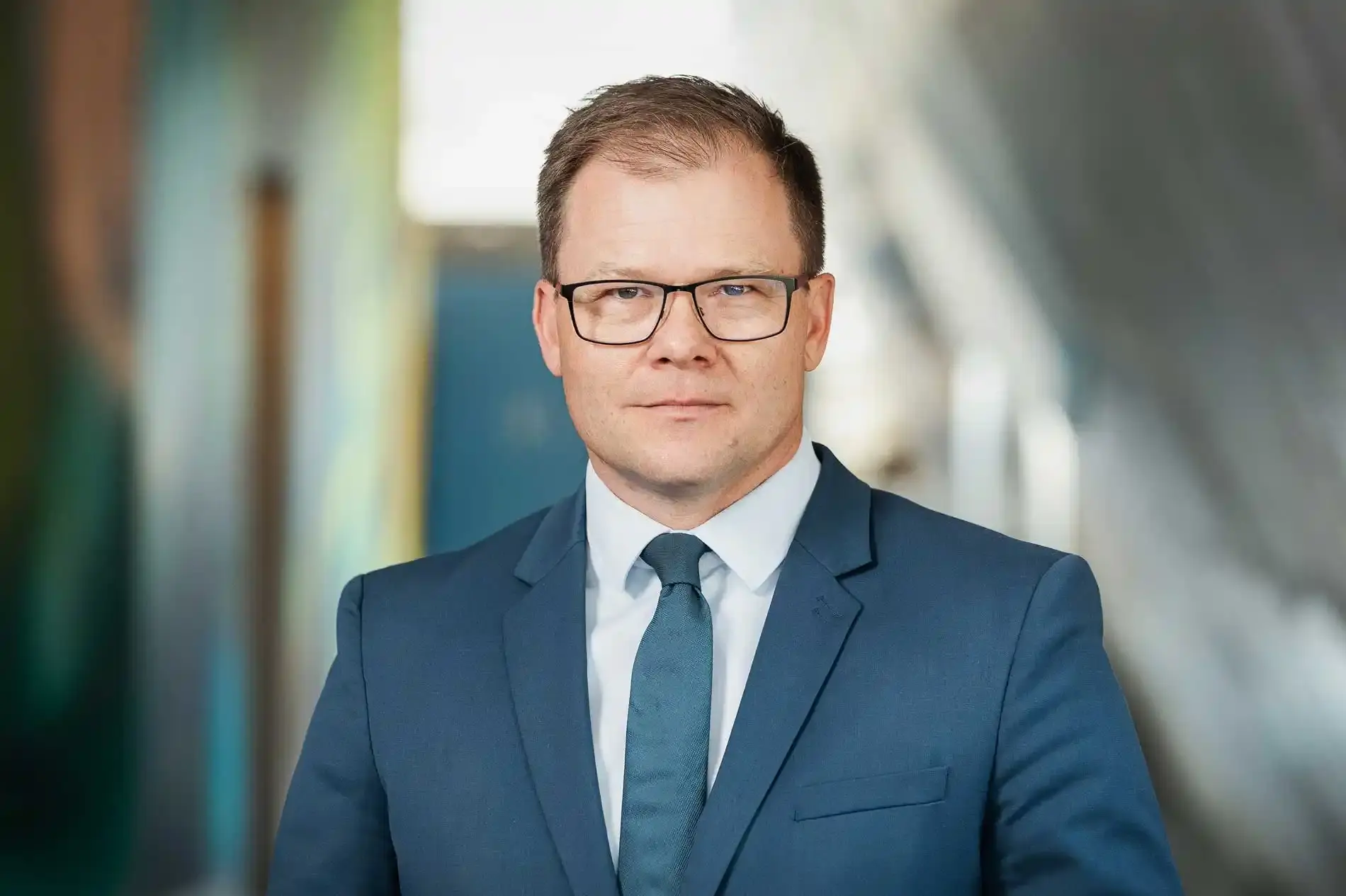 Portrait eines Mannes mit kurzen braunen Haaren und dunkler Brille. Er trägt einen blauen Anzug und Krawatte. Der Hintergrund ist unscharf.