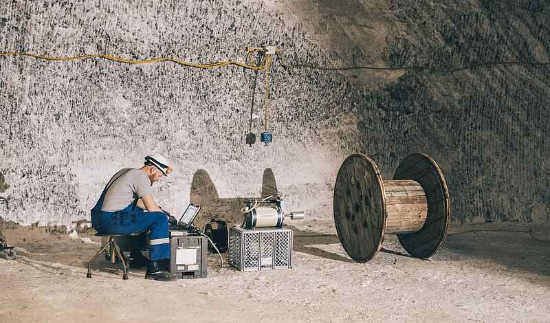 Ein Mann in Arbeitsbekleidung und Helm sitzt an einem Notebook. Vor ihm ist ein Messinstrument aufgebaut, das auf eine hölzerne Kabeltrommel gerichtet ist.