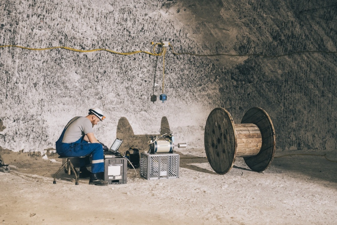 Ein Mann in Arbeitsbekleidung und Helm sitzt an einem Notebook. Vor ihm ist ein Messinstrument aufgebaut, das auf eine hölzerne Kabeltrommel gerichtet ist.