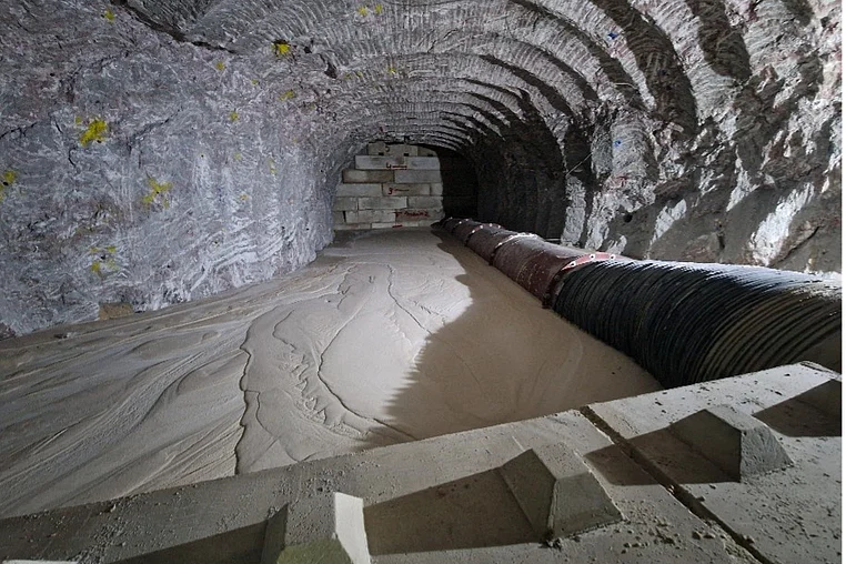 •	Das Bild zeigt einen Streckenbereich im Bergwerk. Im Hintergrund liegt das eine Ende des Streckenbereichs. Dort befindet sich eine Mauer aus grauen Blocksteinen. Im Vordergrund ist das andere Ende des Streckenbereichs. Hier befindet sich eine weitere Mauer im Anschnitt. Im dazwischenliegenden Bereich ist flüssiger Beton zu erkennen. Rechts neben dem Beton ist der obere Teil einer schwarzen Röhre mit Rillen zu erkennen. Der umliegende Bereich, also die Decke und Wände zeigen ein wellenartiges Muster im Salz. 