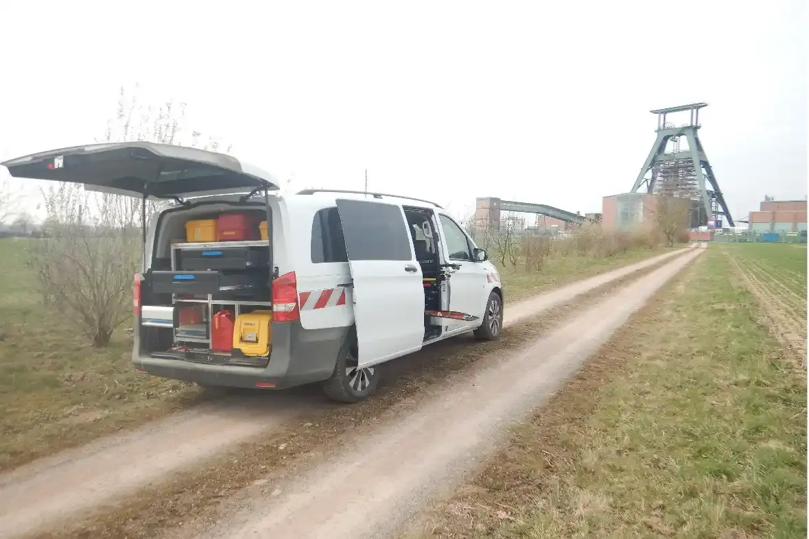 Ein weißer Kastenwagen steht mit geöffneter Heckklappe und geöffneter Seitenschiebetür auf einem Feldweg. Im Hintergrund ist eine Industrieanlage mit Förderturm zu sehen.