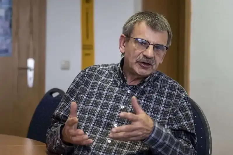 Foto eines Mannes mit braun-grauen Haaren und Bart im karierten Hemd. Der Mann sitzt an einem Tisch und erzählt und gestikuliert mit den Händen.