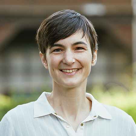 Portrait einer Frau mit kurzen dunklen Haaren und hellem Oberteil mit Kragen, die in die Kamera lächelt. Der Hintergrund ist unscharf.