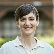 Portrait einer Frau mit kurzen dunklen Haaren und hellem Oberteil mit Kragen, die in die Kamera lächelt. Der Hintergrund ist unscharf.