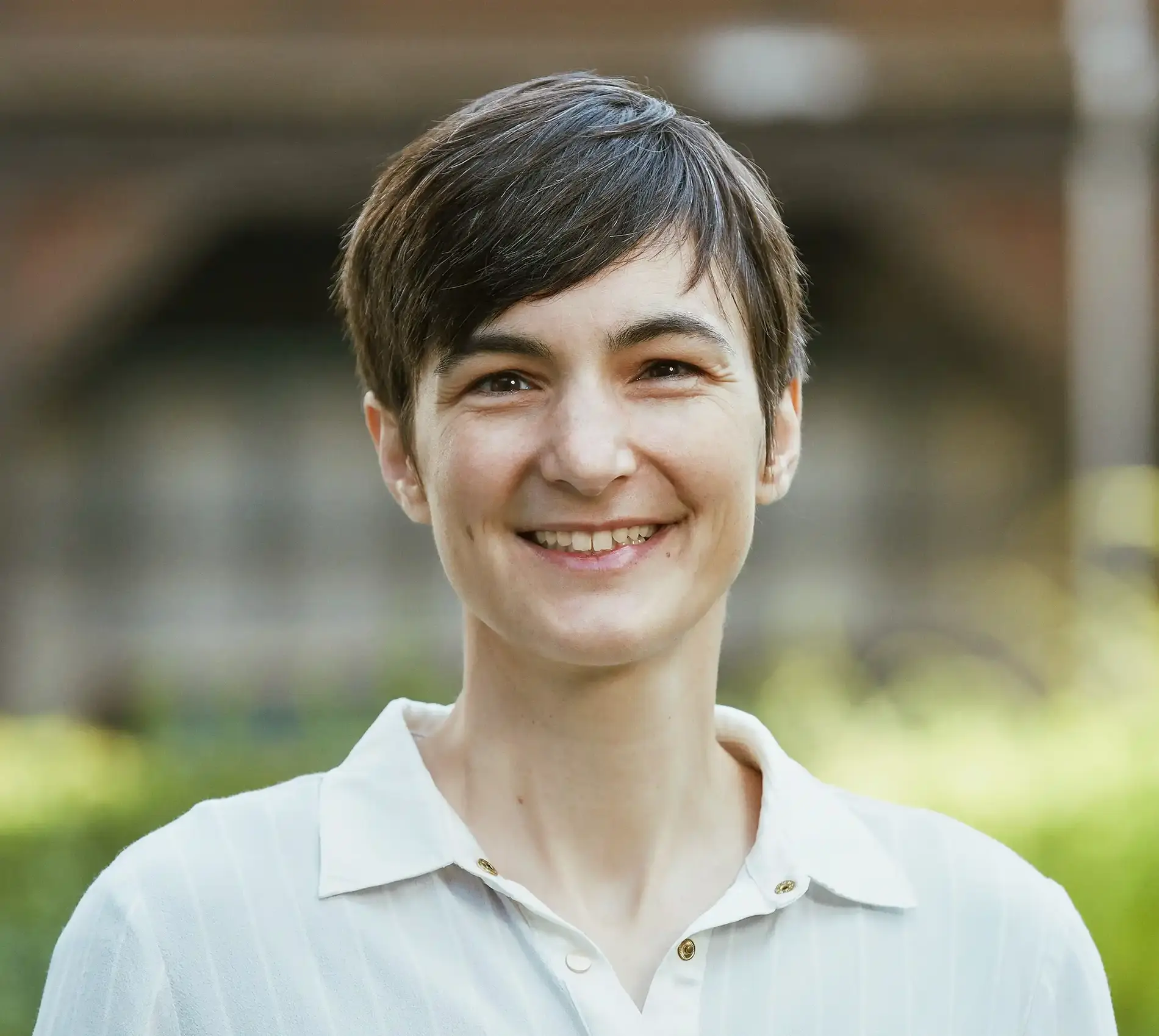 Portrait einer Frau mit kurzen dunklen Haaren und hellem Oberteil mit Kragen, die in die Kamera lächelt. Der Hintergrund ist unscharf.