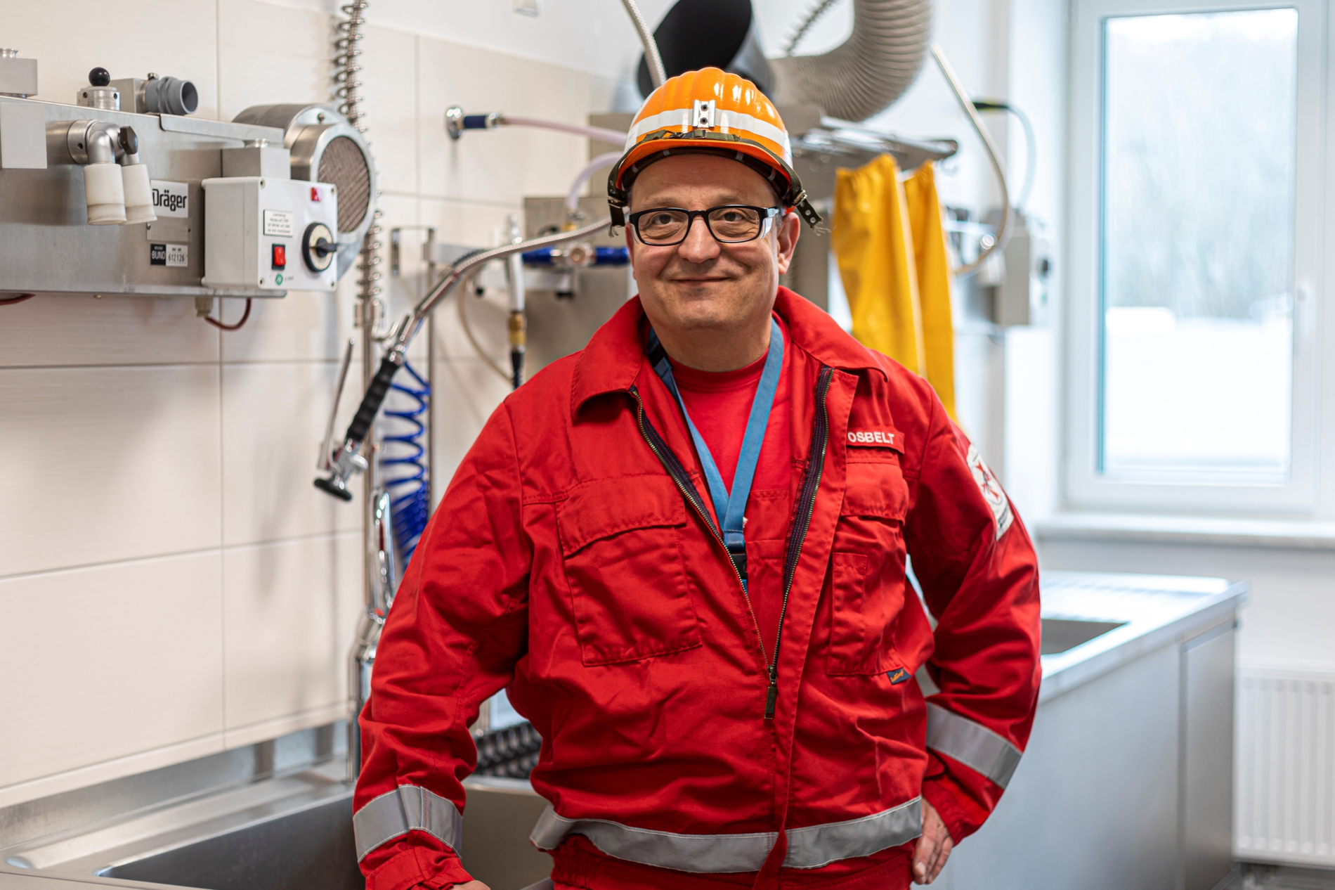 In einem Labor steht ein Mann vor einer gefliesten Wand. Er trägt einen Schutzhelm und eine Brille und lächelt in die Kamera.