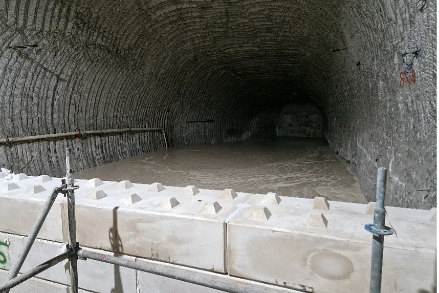 Das Bild zeigt einen Streckenbereich im Bergwerk. Die Strecke wird im Hintergrund durch eine Wand begrenzt. Im Vordergrund ist eine Mauer aus Beton-Blocksteinen und Teile eines Gerüsts zu erkennen. Der dazwischenliegende Bereich wird über eine an der linken Streckenwand montierten Rohrleitung mit flüssigem Beton verfüllt.