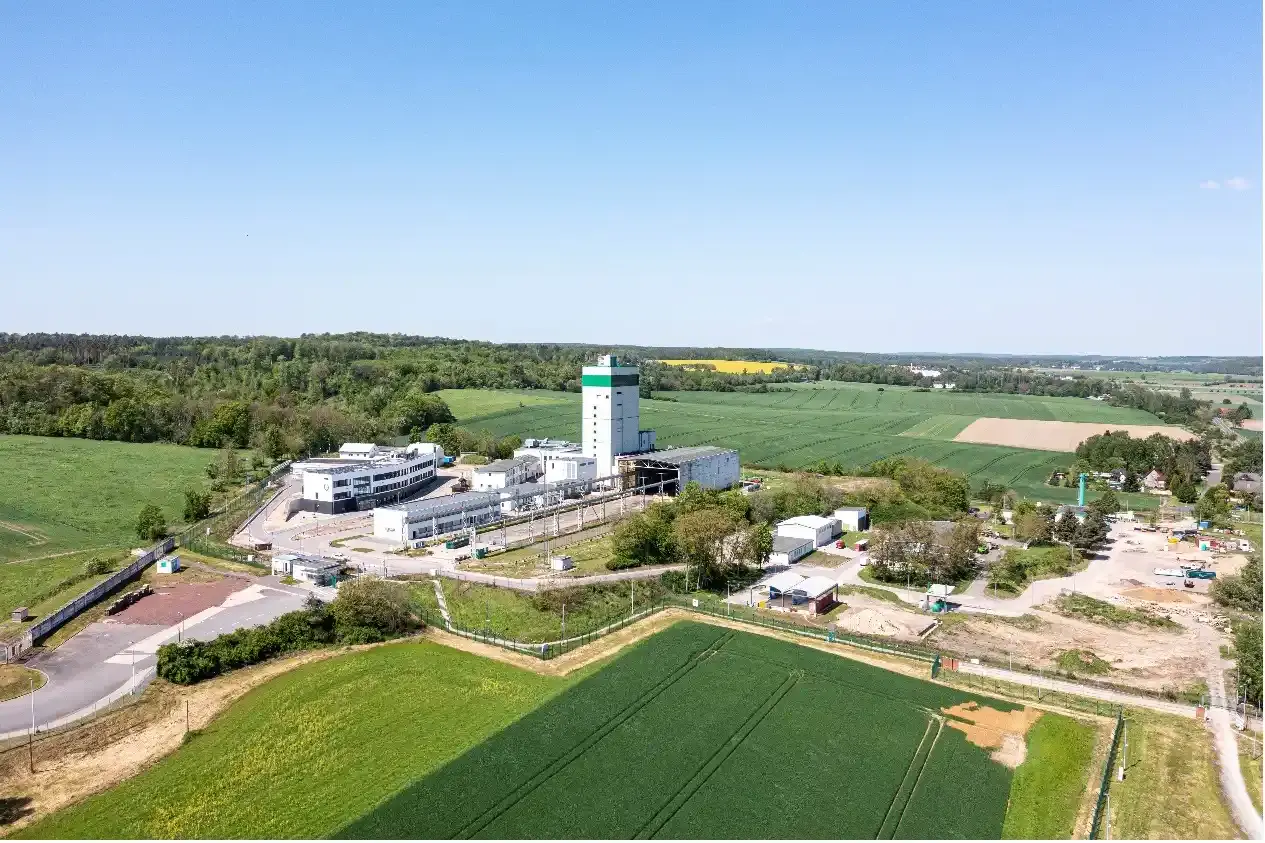 Luftaufnahme eines Industriegeländes. Mittig steht ein grün-weißer Turm. Das Gelände ist umgeben von Feldern und Wald.