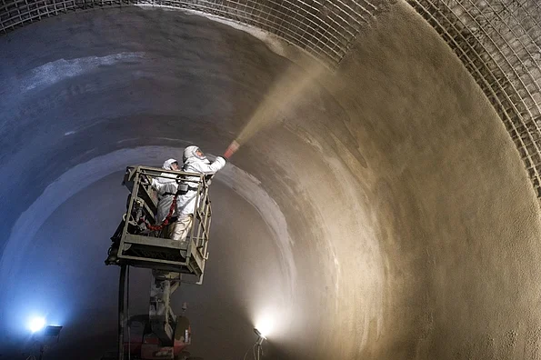 Zwei Arbeiter*innen in weißen Schutzanzügen stehen auf einer Hubbühne und spritzen Beton aus einem Schlauch auf eine Wandfläche.