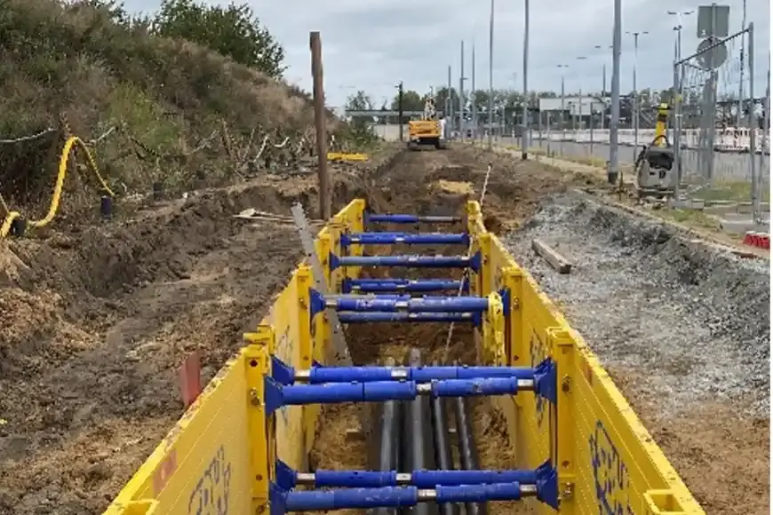 Rohre werden in einer Baugrube verlegt. Gelbe Spundwände und blaue Stützen stabilisieren die Baugrube.