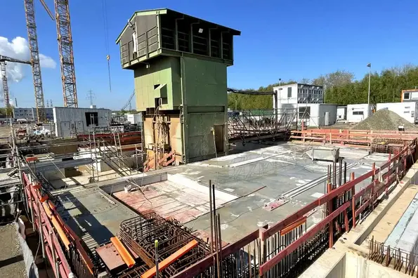 Ein grünes Haus – die temporäre Fördereinrichtung – steht im Umfeld einer Baustelle. Eine Betondecke ist um das Gebäude errichtet.