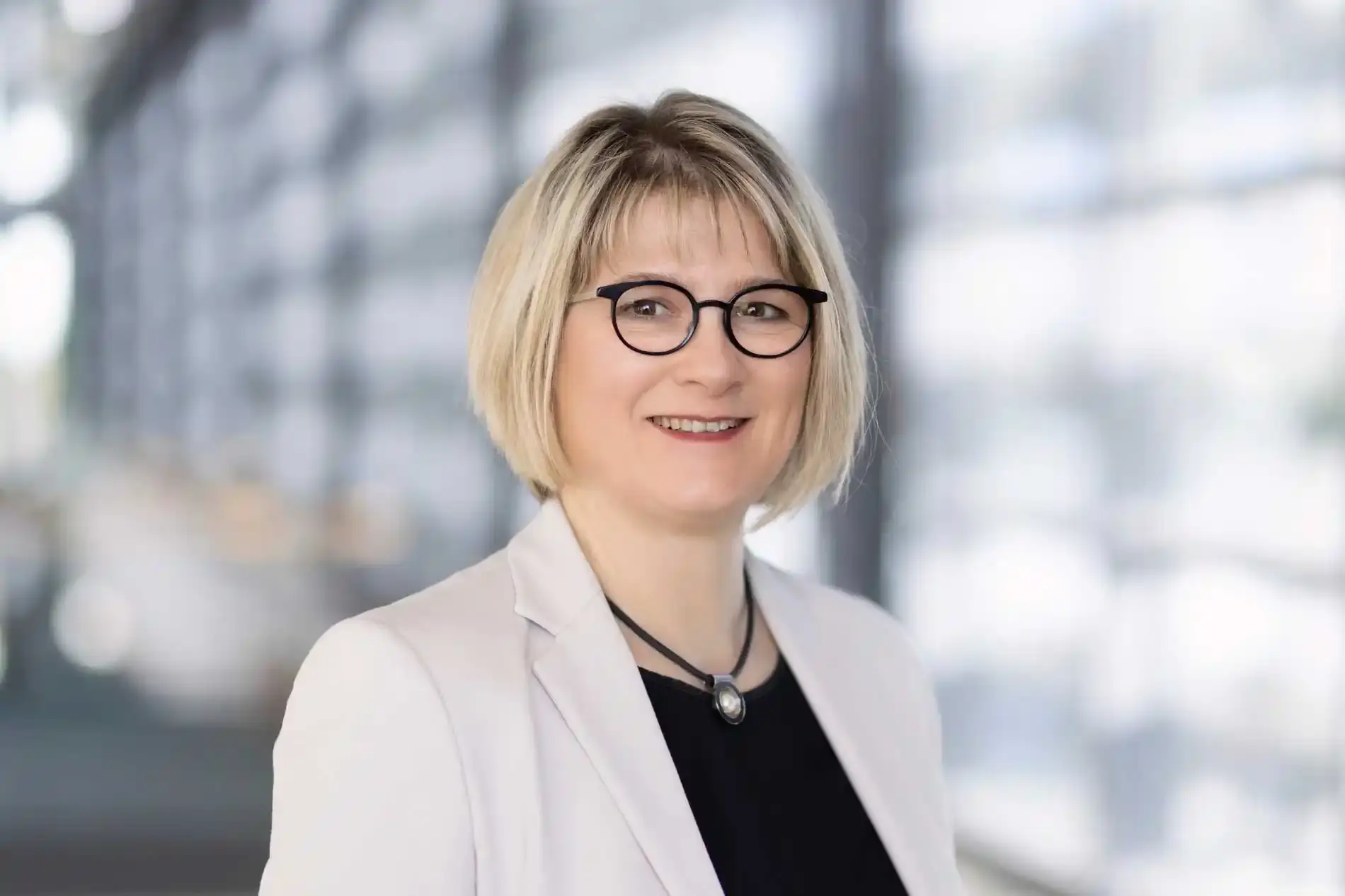 Portrait photo: A woman with blond chin-length hair, black glasses and a light-colored blazer looks friendly into the camera