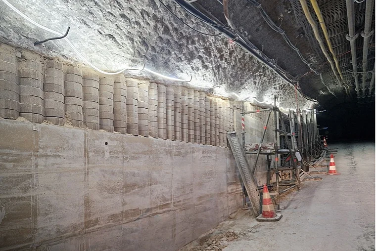 Das Bild zeigt einen ansteigenden Streckenbereich im Bergwerk. Unterhalb des Deckenbereichs mit typischen Frässpuren im Salz erstreckt sich auf der linken Seite eine neu errichtete Wand aus Beton bis zum Boden. Das obere Drittel der Betonwand besteht aus aufeinander gebauten Rasengittersteinen, der untere Bereich aus einer glatten Betonschicht. Im oberen Streckenverlauf ist Wand noch mit Schalungswänden und Metallstützen versehen, hinter denen der frische Beton aushärtet.