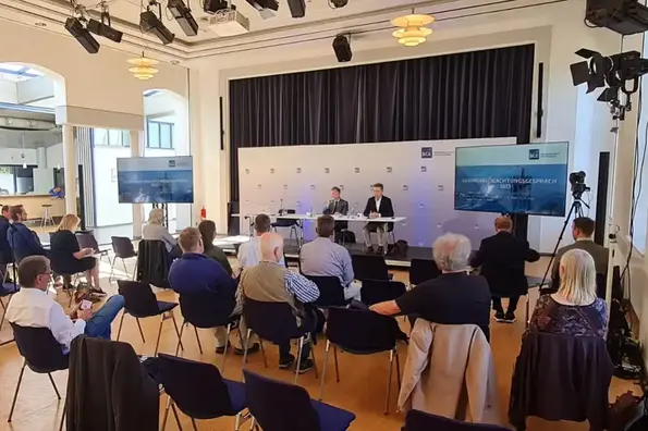 Das Foto zeigt den Blick in den Veranstaltungsraum. Personen sitzen in mehreren Reihen auf Stühlen und blicken in Richtung Podium. Am Podium sitzen BGE-Mitarbeiter und halten einen Vortrag. Auf zwei Monitoren werden die Präsentationen abgebildet.