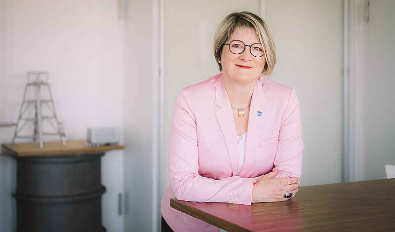 Frau in weißer Bluse und rosafarbenem Blazer lehnt an einem hohen Bürotisch. Sie lächelt in die Kamera, hat blonde halblange Haare und trägt eine Brille mit dunklem Rahmen. Im Hintergrund sind unscharf weiße Wände und ein grauer Stehtisch zu sehen.