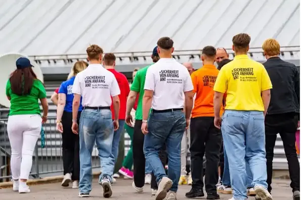 Das Foto zeigt die Rückansicht einer Gruppe junger Menschen in bunten T-Shirts mit der Aufschrift „Sicherheit von Anfang an! Azubi-Wettbewerb BG RCI 2024/2025“