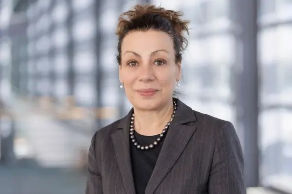 Porträtfoto: Eine Frau mit hochgebundenem brauen Haar, dunkler Perlenkette und schwarzer Bluse blickt freundlich in die Kamera