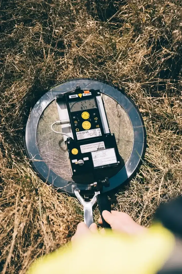 Blick von oben auf Strahlungsmessgerät, das auf begrastem Boden aufliegt und von zwei Händen festgehalten wird