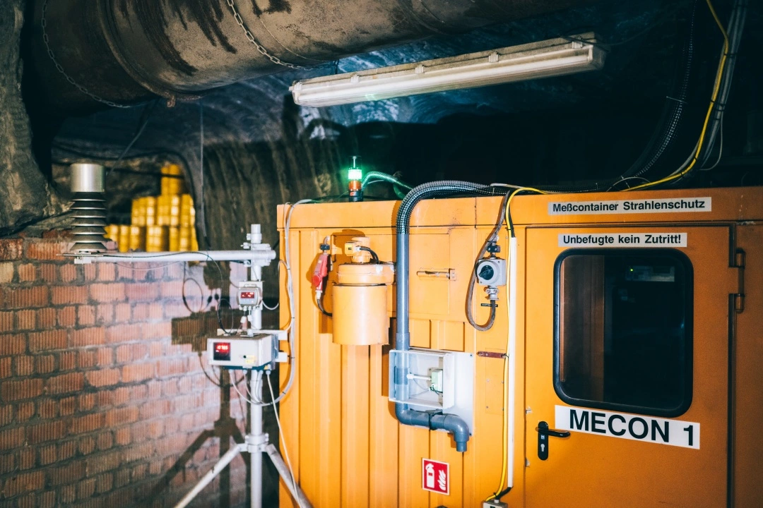 Ein galber Container steht neben einer gemauerten Wand. Davor stehen Messgeräte. An dem Container hängen Hinweisschilder und andere Instrumente.