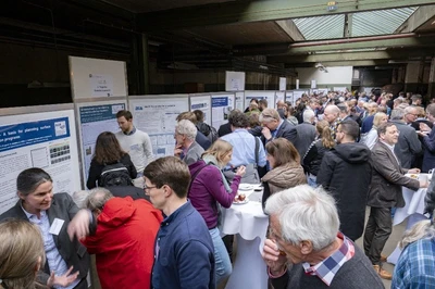 Zu sehen sind zahlreiche Menschen zu sehen, die sich Poster mit wissenschaftlichen Inhalten ansehen und darüber diskutieren.