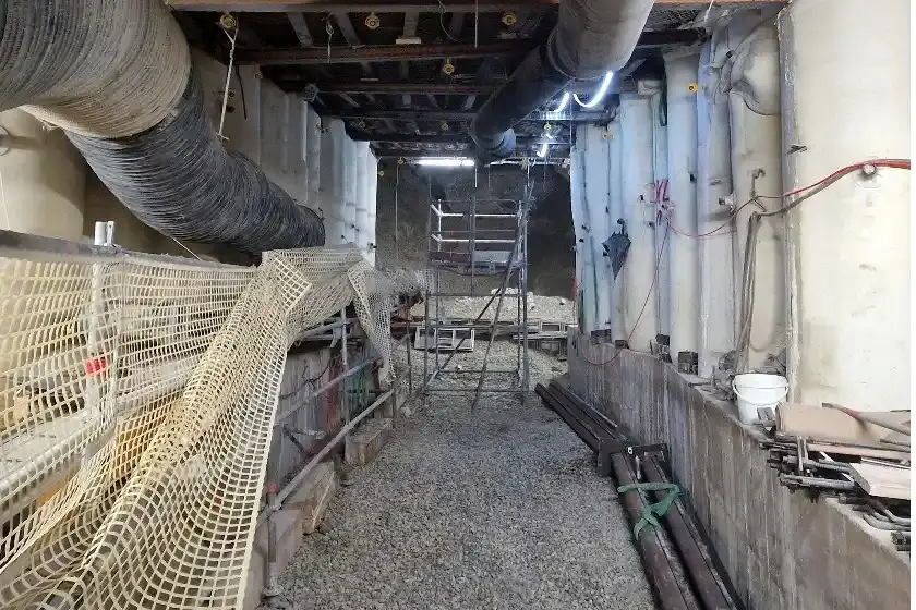 Das Bild zeigt den Blick in einen Abbau im Bergwerk. Auf der linken und rechten Seite stehen helle, säulenartige Stützbauwerke auf einem grauen Betonfundament. Quer über die Stützbauwerke, direkt unterhalb der Decke, verlaufen dunkle Metallträger. Unterhalb der Metallträger verlaufen zwei dicke, schwarze Schläuche. Zwischen den Stützbauwerken steht ein Gerüst. Im Hintergrund und auf dem Boden liegt Schotter.