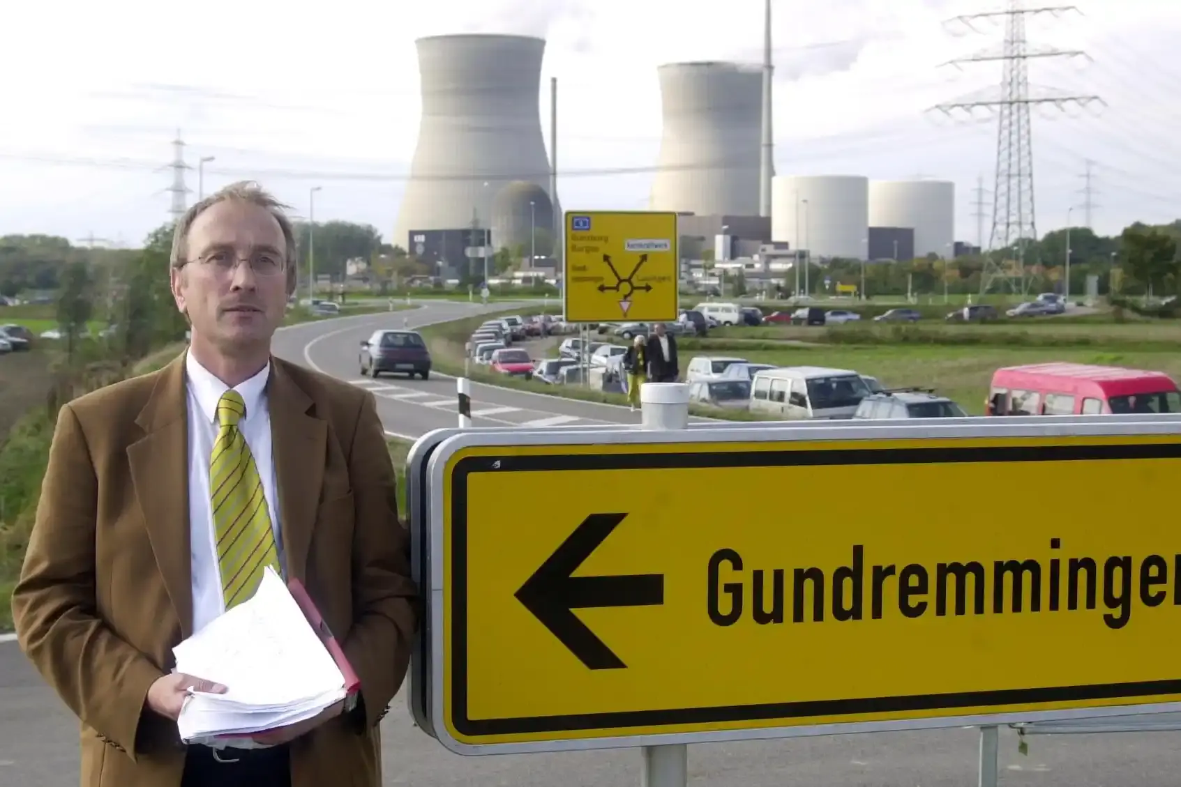 Ein Mann in Anzug steht vor Kühltürmen eines Atomkraftwerkes. In der Hand hält er mehrere Papiere. Zwischen ihm und den Kühltürmen ist eine Autobahn zu sehen. Neben ihm steht das Ortsschild Grundremmngen.