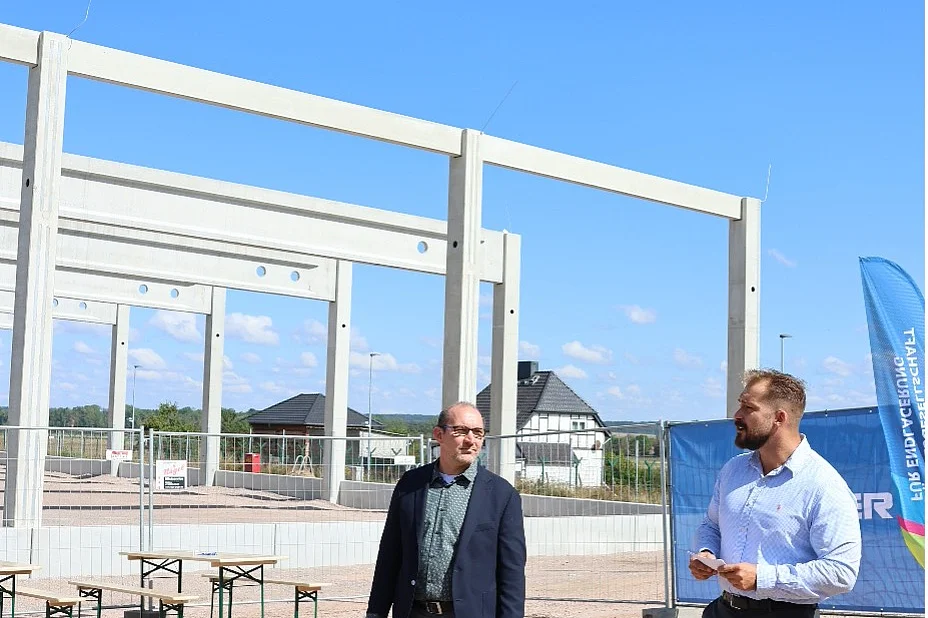 Das Bild zeigt eine Baustelle mit weißen Betonstützen vor einem blauen Himmel. Im Vordergrund stehen zwei Personen.