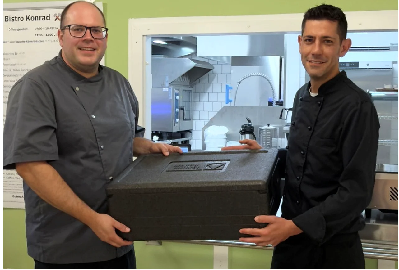 Zwei Köche halten eine schwarze Box mit Deckel. In dieser Box sind bestellte Brötchen und Baguettes für die Bergleute unter Tage. 