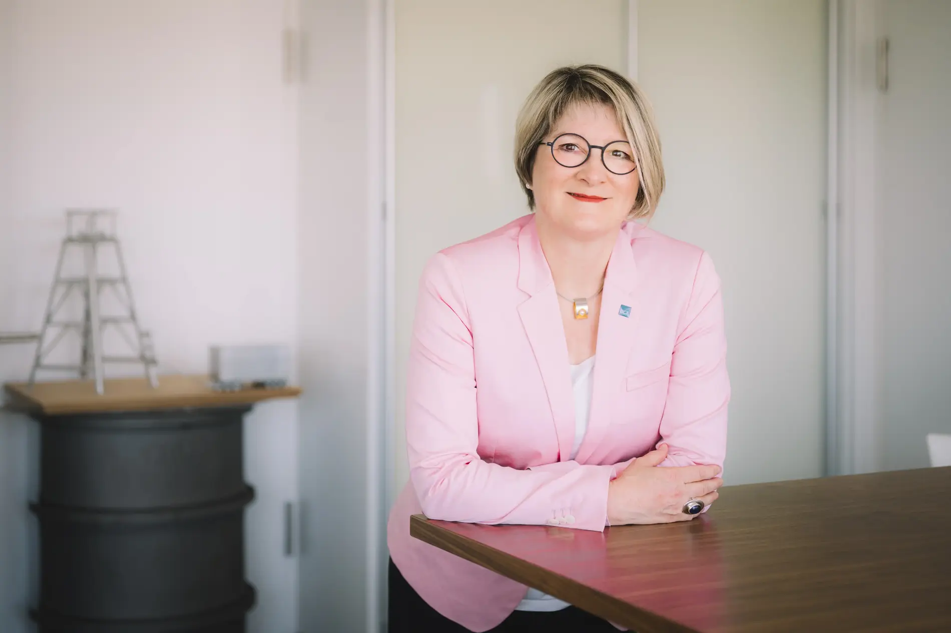 Frau in weißer Bluse und rosafarbenem Blazer lehnt an einem hohen Bürotisch. Sie lächelt in die Kamera, hat blonde halblange Haare und trägt eine Brille mit dunklem Rahmen. Im Hintergrund sind unscharf weiße Wände und ein grauer Stehtisch zu sehen.