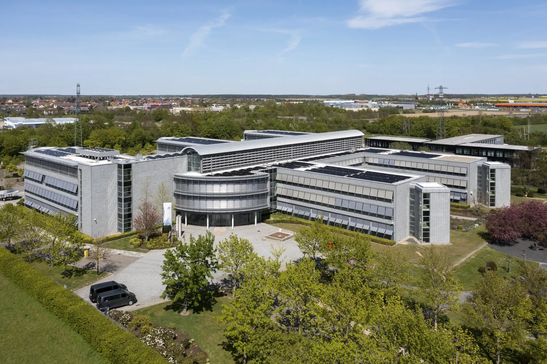 Aerial view of a gray-silver office building. Trees, residential buildings and blue sky can be seen in the background. 