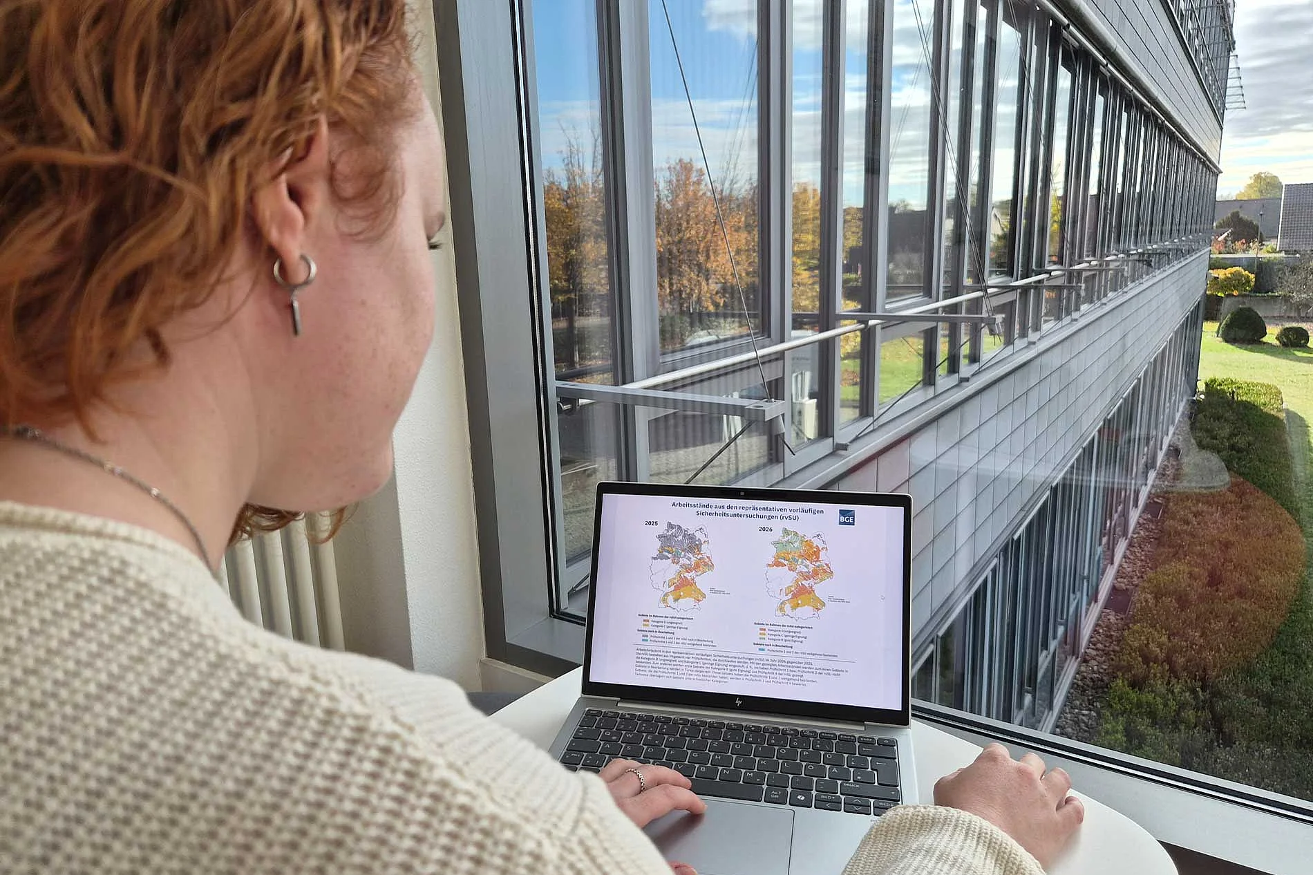[Translate to English:] Over-the-shoulder view: A woman with red, curly hair is looking at a screen displaying two maps of Germany.