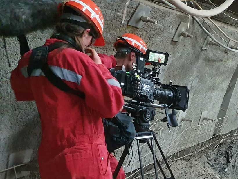 Zwei Kameraleute in rotem Overall und mit rotem Schutzhelm filmen in einem Bergwerk.