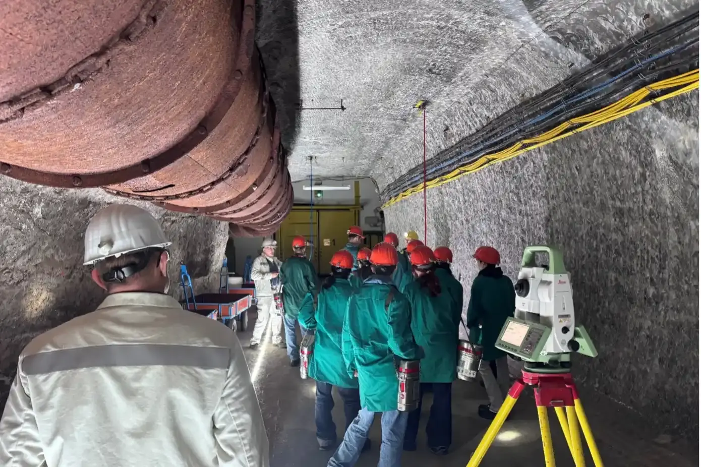 Eine Gruppe von Personen mit Schutzhelmen und Bergwerkskleidung in einem Tunnel. An den Wänden und an der Decke verlaufen Rohre.