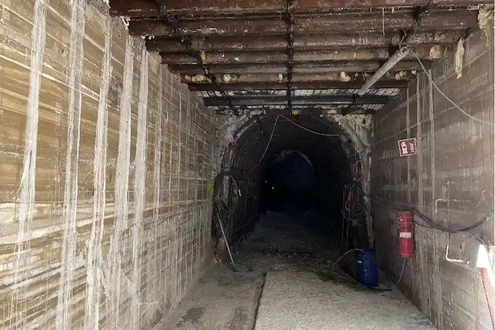Das Bild zeigt den Blick in eine Strecke im Bergwerk. Über die ersten Meter erstrecken sich graue Betonwände an beiden Seiten der Strecke. Quer auf den Betonwänden sind mehrere Stahlträger verbaut, die die Wände verbinden. Im hinteren Teil schließt sich ein Bereich an, in dem die Strecke durch mehrere, hintereinander verbaute, halbrunde Stahlbögen gestützt wird.